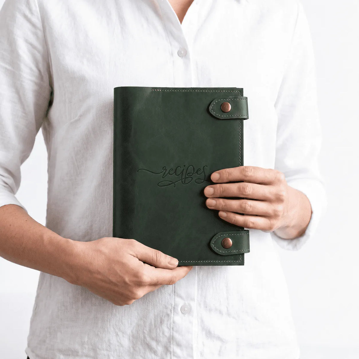 A person in a white linen shirt holding a dark green leather recipe book with 'recipes' written in cursive on the front cover.