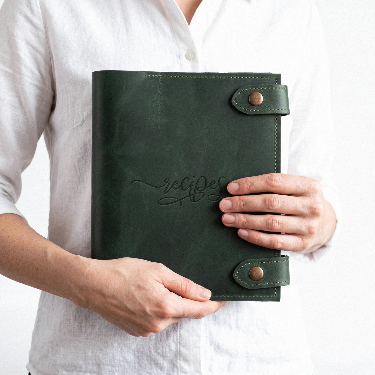 A person in a white linen shirt holds a dark green leather recipe book with 'recipes' embossed on the cover, secured by metal snap closures.