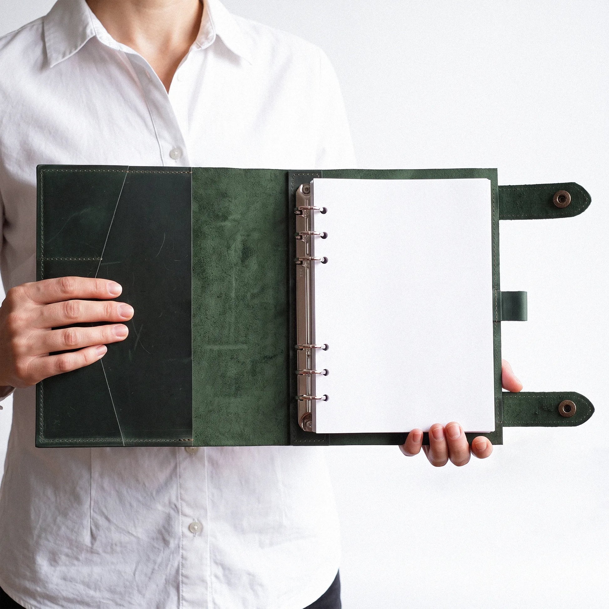 A person in a white button-up shirt holds open a dark green leather ring binder notebook with blank white pages against a plain white background.