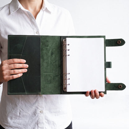 A person in a white button-up shirt holds open a dark green leather ring binder notebook with blank white pages against a plain white background.