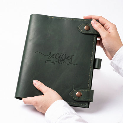 A dark green leather recipe binder with 'recipes' embossed on the front, held by two hands against a clean white background.
