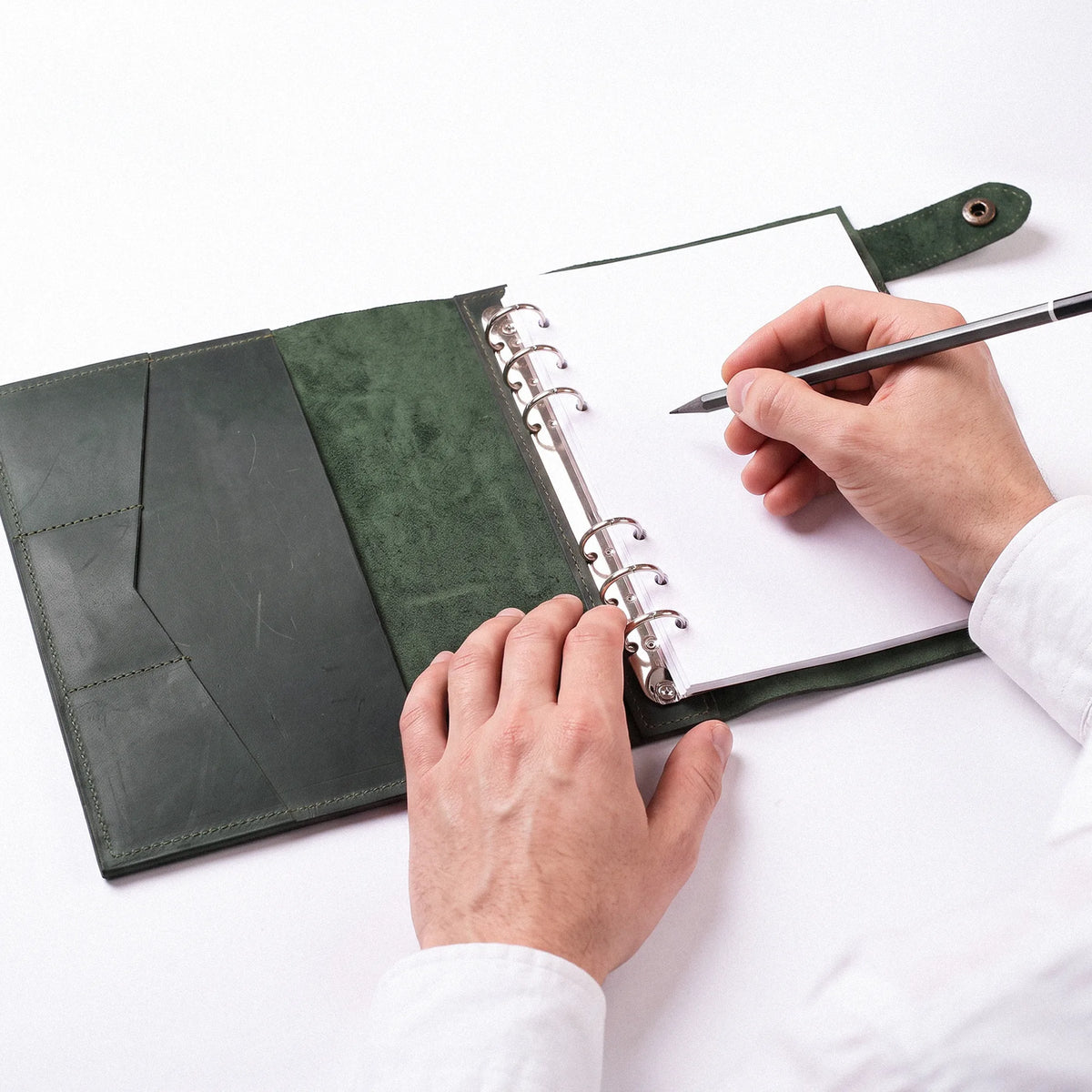 A top-down view of a person's hands using a black pen to write in a green leather-bound ring binder notebook on a clean white surface.