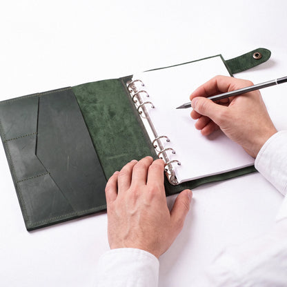 A top-down view of a person's hands using a black pen to write in a green leather-bound ring binder notebook on a clean white surface.